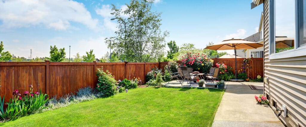 Jardin arrière aménagé avec pelouse, clôture en bois, et terrasse dallée. Mobilier de jardin, parasol et rosiers colorés créent un espace détente.